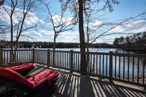 A serene lakeside view from a deck with red lounge chairs, surrounded by trees and a clear blue sky.
