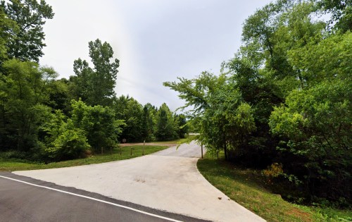 A paved road curves into a wooded area with lush green trees on either side under a cloudy sky.
