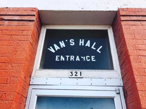 Sign above a door reading "Van's Hall Entrance" with the number 321 below it, framed by red brick.