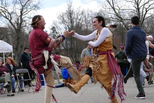 Two dancers in traditional attire perform a lively dance outdoors, surrounded by spectators and trees.