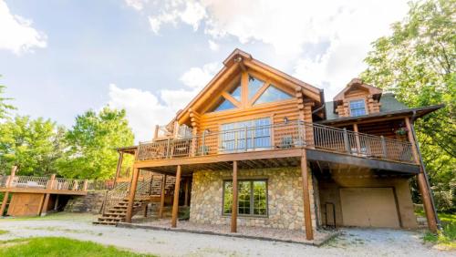 A large log cabin with a stone base, featuring a spacious deck and surrounded by greenery under a blue sky.