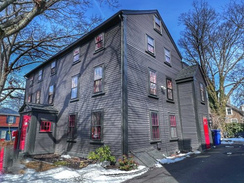 A large, dark-colored house with red windows and doors, surrounded by snow and bare trees under a clear blue sky.