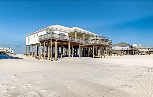 A beach house on stilts overlooking a sandy shore under a clear blue sky.