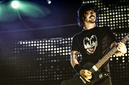 A musician performs on stage, wearing a black shirt with a graphic design and holding an electric guitar.