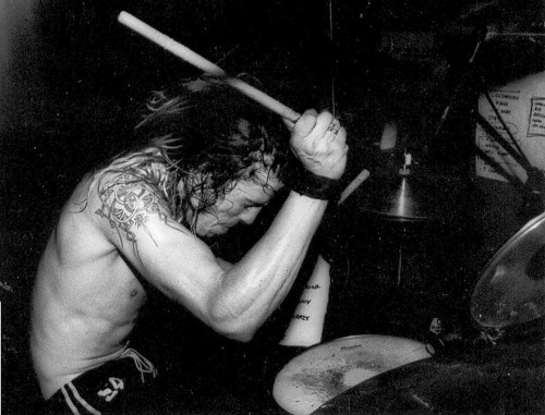 A shirtless drummer passionately playing the drums, with long hair and tattoos visible on his arms.