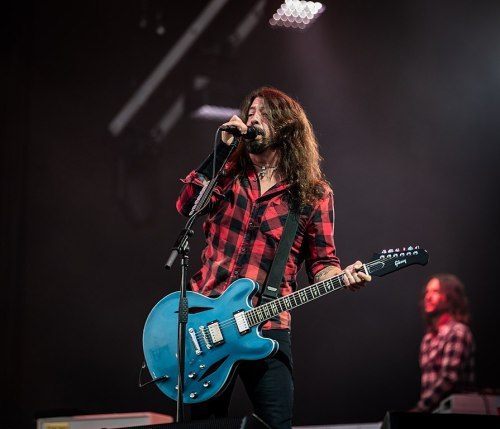 A musician with long hair sings into a microphone while playing a blue guitar on stage, wearing a red plaid shirt.