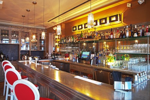 A stylish bar with a polished counter, shelves of liquor, and elegant lighting, featuring red and white bar stools.