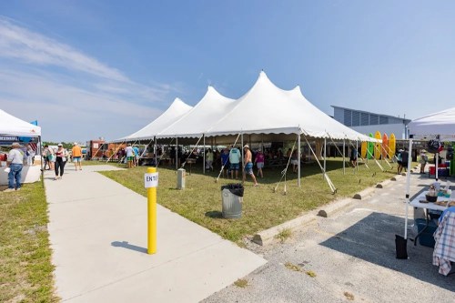 A large white tent at an outdoor event, with people walking and various booths set up nearby.