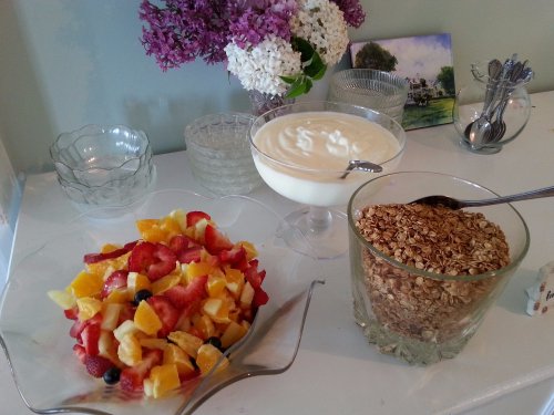 A table with a fruit salad, yogurt, and a glass container of granola, surrounded by flowers and decorative bowls.