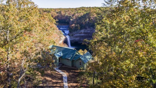 A green house surrounded by trees, overlooking a waterfall and a river in a scenic autumn landscape.