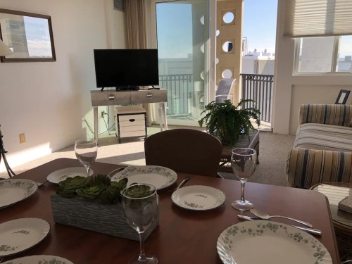A bright living space with a dining table set, a TV, and a balcony view, featuring plants and a cozy sofa.