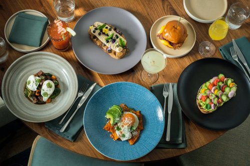 A table set with various colorful dishes, including avocado toast, a burger, and a salad, surrounded by drinks.