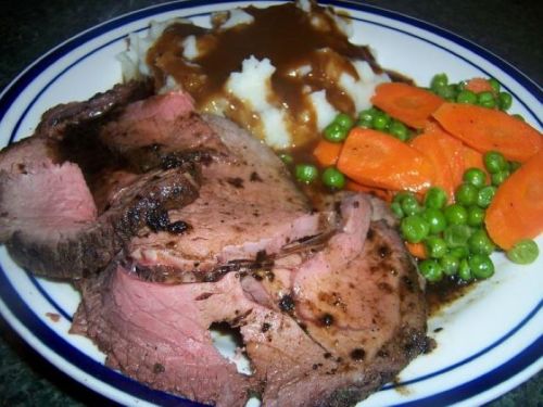 A plate of sliced roast beef with gravy, mashed potatoes, and a side of carrots and peas.