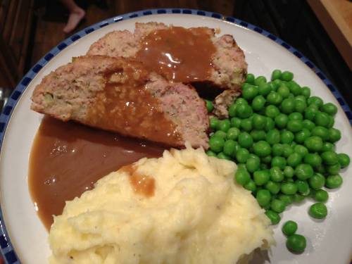 A plate with meatloaf slices topped with gravy, served with mashed potatoes and a side of green peas.