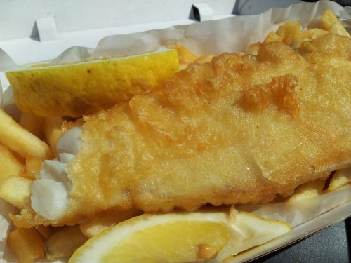 A serving of golden-battered fish with fries and lemon wedges in a takeout container.