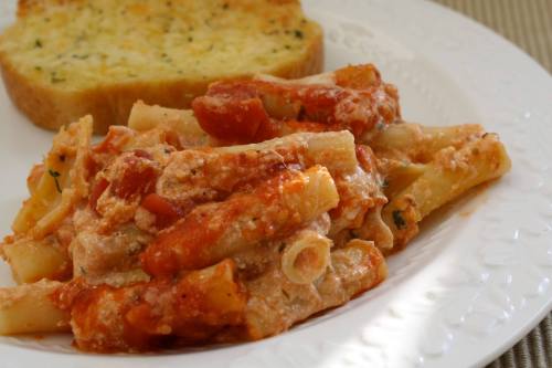 A plate of baked pasta with tomato sauce and cheese, served alongside a slice of garlic bread.