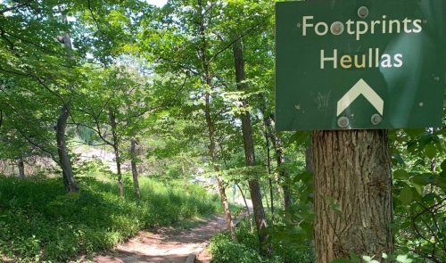 A green trail sign reading "Footprints Heullas" next to a dirt path surrounded by lush greenery.