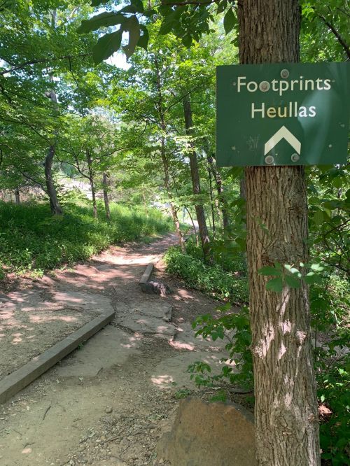 A wooded trail with a sign reading "Footprints Heullas" beside a dirt path surrounded by greenery.