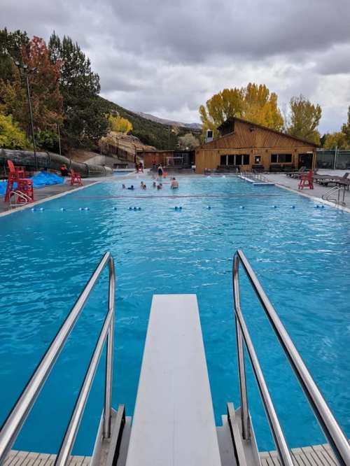 A clear blue swimming pool with a diving board, surrounded by trees and a building in the background under a cloudy sky.