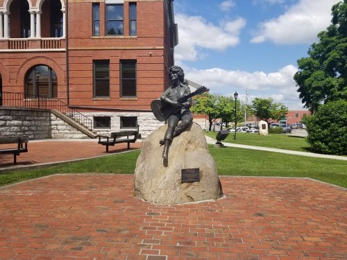A statue of a seated musician with a guitar on a rock, set in a park near a historic building.
