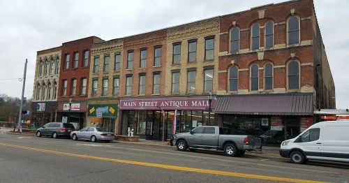Historic brick buildings line a street, featuring shops like "Main Street Antique Mall" and parked vehicles.