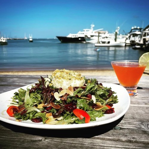 A vibrant salad with greens, almonds, and cherry tomatoes, paired with a cocktail, set against a marina backdrop.