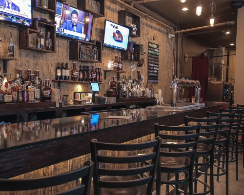 A dimly lit bar with a long counter, shelves of liquor, and two TVs displaying sports. Bar stools line the counter.
