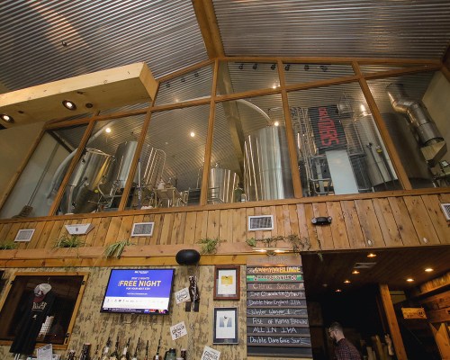 Interior of a brewery with large metal fermentation tanks visible behind a glass wall, wooden decor, and a TV screen.