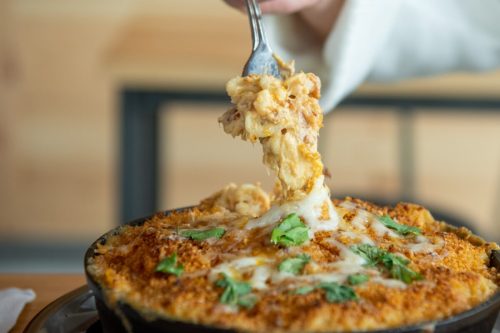 A fork lifts a cheesy, baked pasta dish from a skillet, topped with breadcrumbs and fresh herbs.