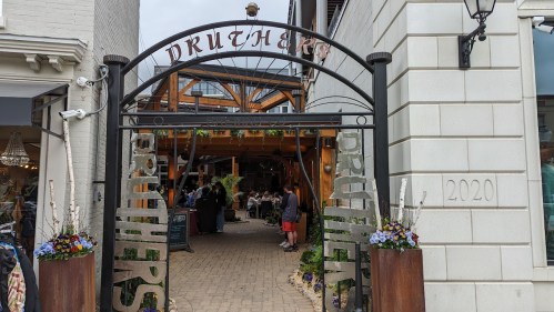 Entrance to a restaurant with a decorative archway, surrounded by plants and people dining outdoors.