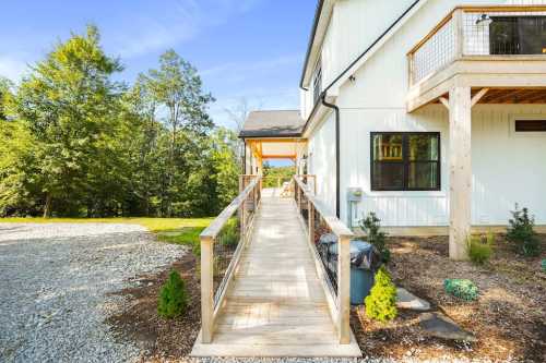 A wooden ramp leads to a modern house surrounded by greenery and gravel, under a clear blue sky.