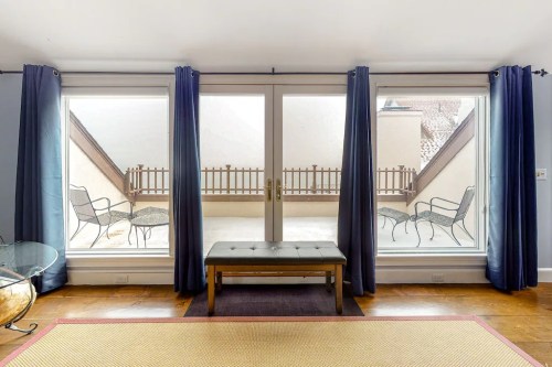 A cozy room with large windows, featuring a bench and two chairs on a balcony outside.