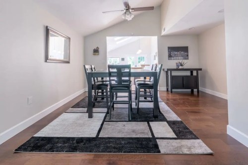 A modern dining area with a wooden table, black chairs, and a patterned rug, featuring natural light from a large window.