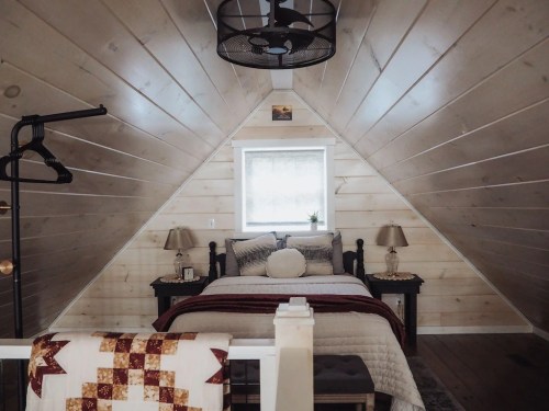 Cozy attic bedroom with sloped wooden walls, a bed, lamps, and a quilt draped over a railing. Natural light from a window.