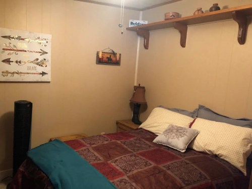 Cozy bedroom with a bed, decorative pillows, a lamp, and wall art in warm tones. Shelves display small decor items.
