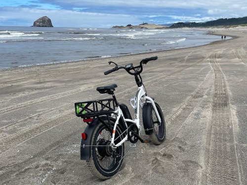 A white electric bike parked on a sandy beach with ocean waves and a distant rocky formation in the background.