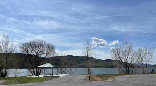 A serene lakeside view with a gazebo, trees, and mountains under a blue sky with scattered clouds.