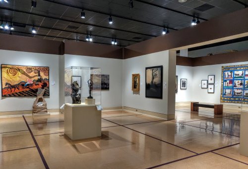 Art gallery interior featuring various artworks, sculptures, and a colorful textile piece on display.