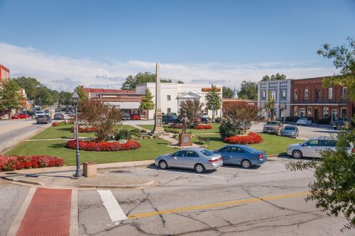A scenic town square with flower beds, cars parked, and buildings under a clear blue sky.