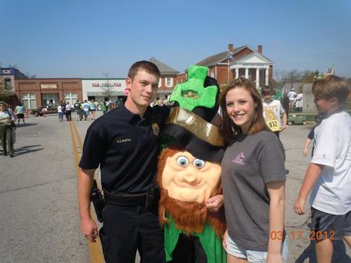 A police officer and a young woman pose with a leprechaun mascot at a St. Patrick's Day event.