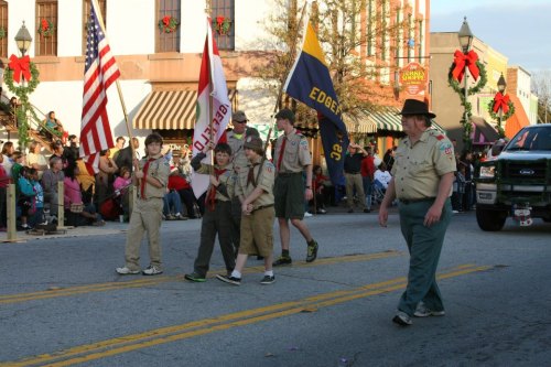Boy Scouts march in a festive parade, carrying flags and walking along a street decorated for the holidays.