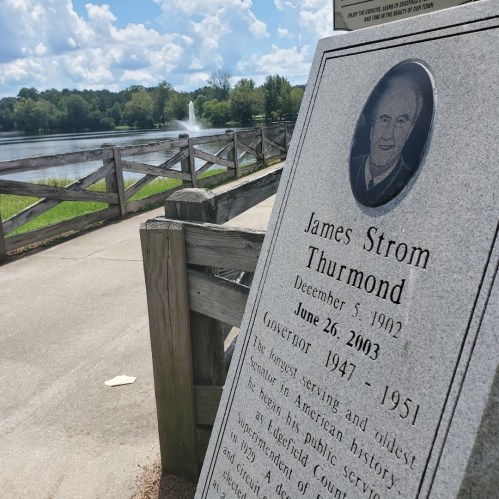 A stone plaque honoring James Strom Thurmond, with a lake and fountain in the background on a sunny day.