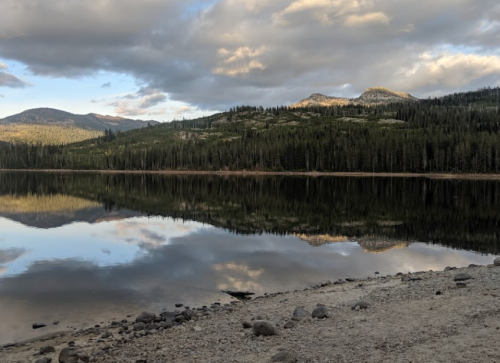 A serene lake reflects mountains and clouds, surrounded by lush forests and a rocky shoreline.