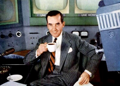 A man in a pinstripe suit sits at a desk, holding a coffee cup, with vintage monitors in the background.