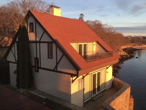 A charming house with a red roof and balcony, overlooking a calm waterway at sunset. Trees line the background.