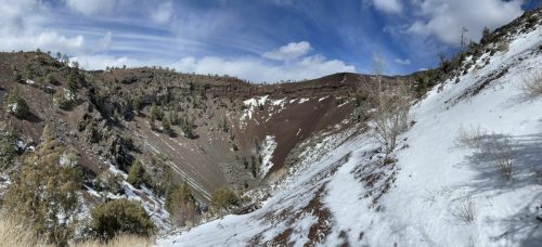 A panoramic view of a snow-covered mountain landscape with rocky slopes and scattered trees under a blue sky.