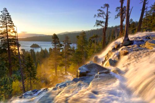 A cascading waterfall flows over rocks, surrounded by trees, with a serene lake and mountains in the background at sunset.