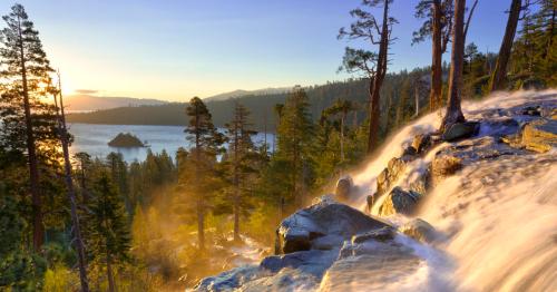 A scenic view of a waterfall cascading over rocks, with a lake and mountains in the background during sunset.