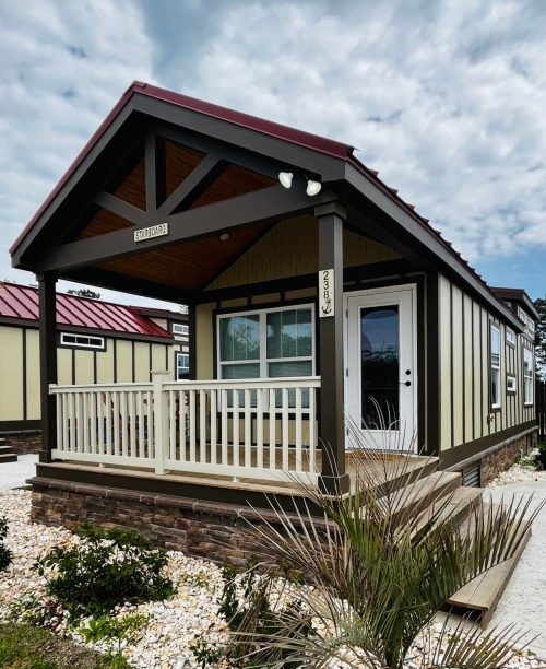 A small, modern house with a porch, featuring a gabled roof and light-colored siding, surrounded by landscaping.