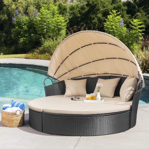 A round outdoor daybed with a beige canopy, cushions, and a basket of towels by a pool surrounded by greenery.